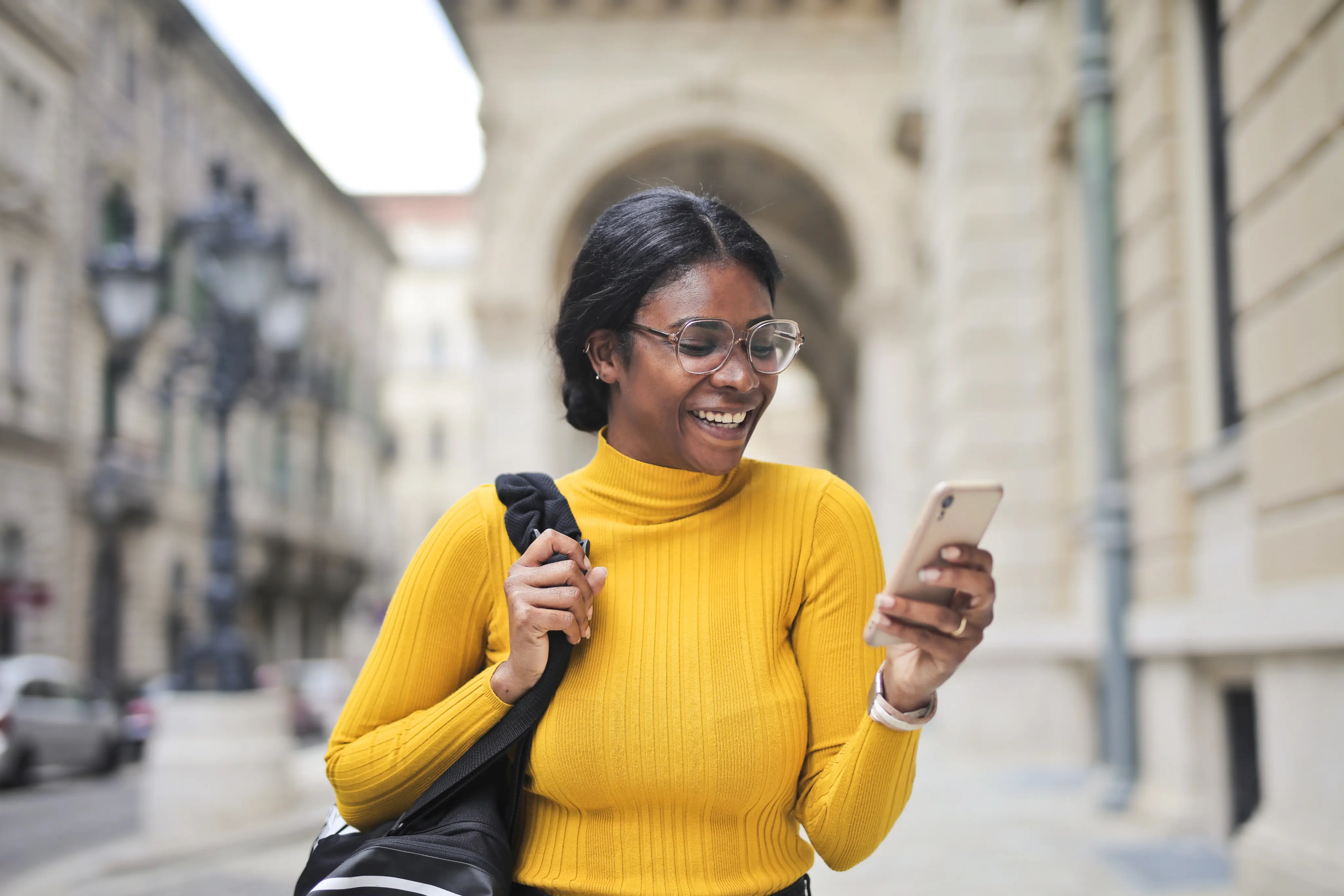 a black woman smiling at her phone with a brown shopping bag over her shoulder